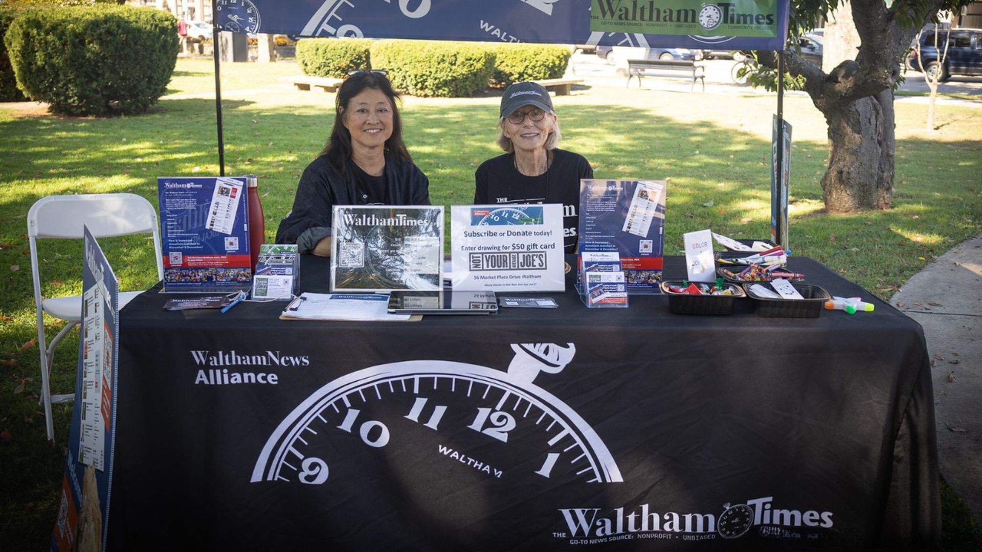 Two women smiling while sitting at a Waltham Times information booth.